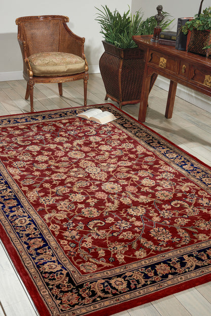 Red oriental rug with blue border in a living room with wicker chair, wooden desk, greenery, and an open book.