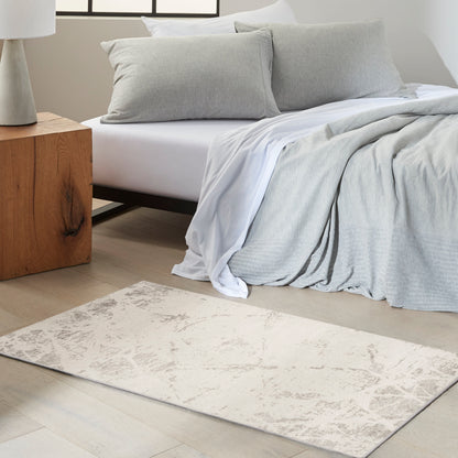 Pale textured area rug on the floor with light gray bedding, white sheets and soft gray pillows, and a wooden nightstand nearby.