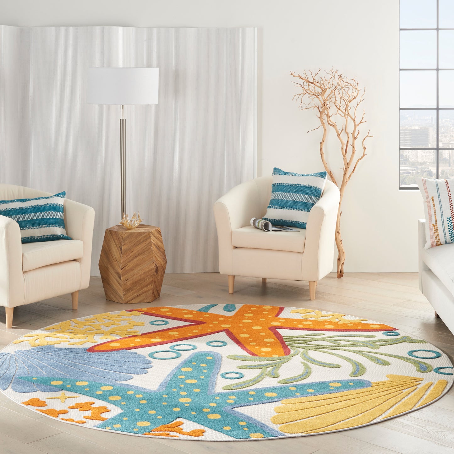 Bright living room with round rug featuring orange and turquoise starfish motif; cream armchairs, teal pillows, wood side table, floor lamp.