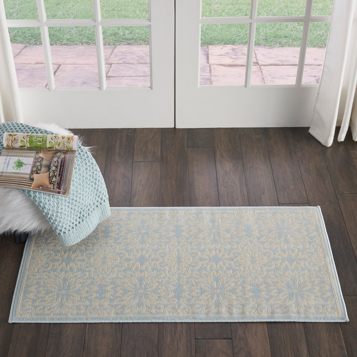 Rectangular light-blue rug with beige floral motif on dark wood floor by French doors; knit throw & magazines nearby.
