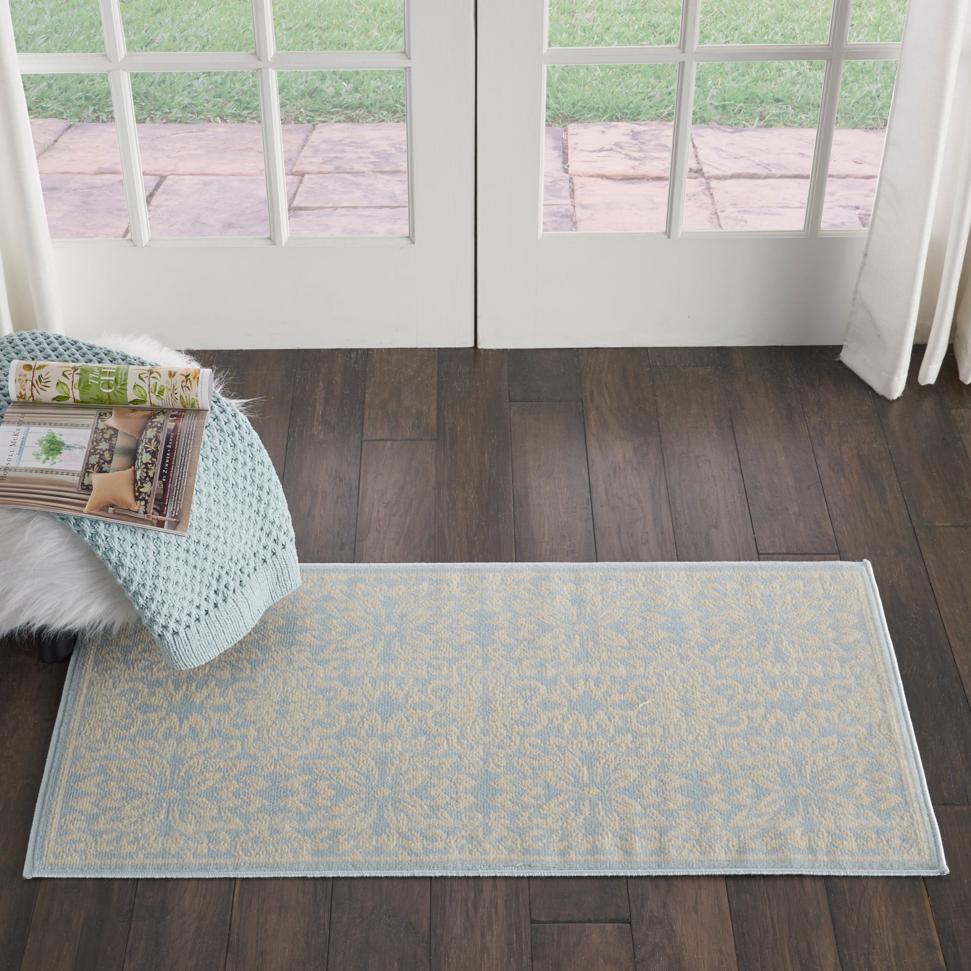 Rectangular light-blue rug with beige floral motif on dark wood floor by French doors; knit throw & magazines nearby.
