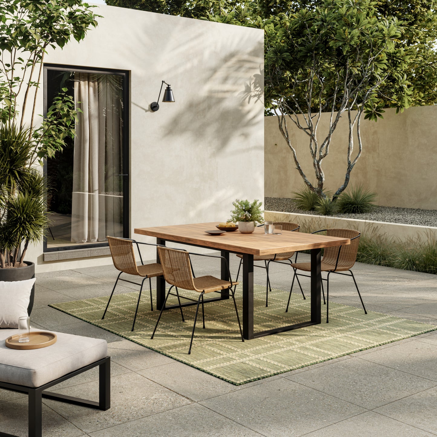 Outdoor dining set: rectangular wood table with black metal legs and wicker chairs on a green rug by a beige stucco wall.