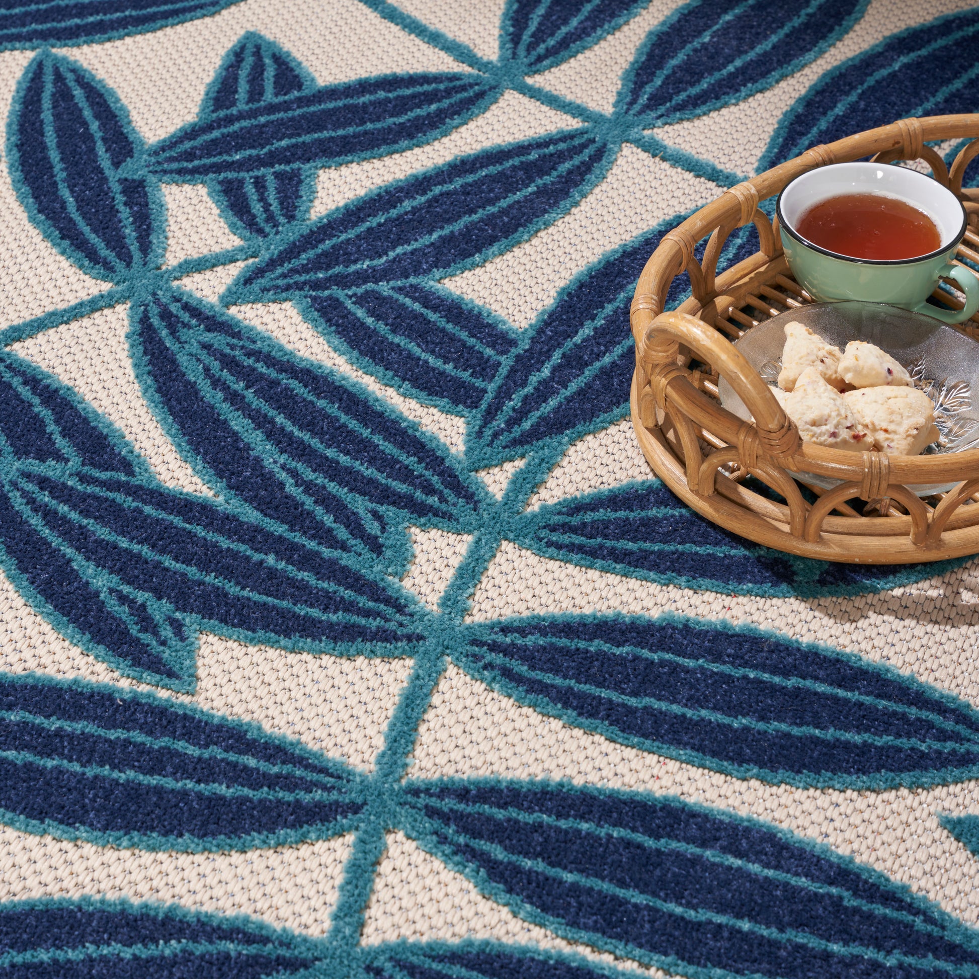 Blue leaf-patterned woven rug with beige background; wicker basket holding a green ceramic cup of tea and cookies.