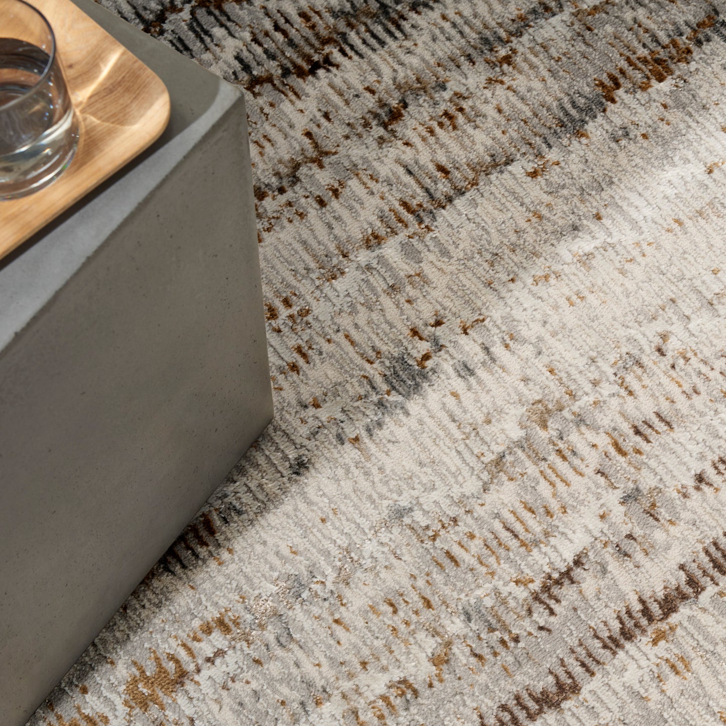 Close-up of a textured beige-gray rug with brown streaks beside a concrete edge and wooden tray with a glass.