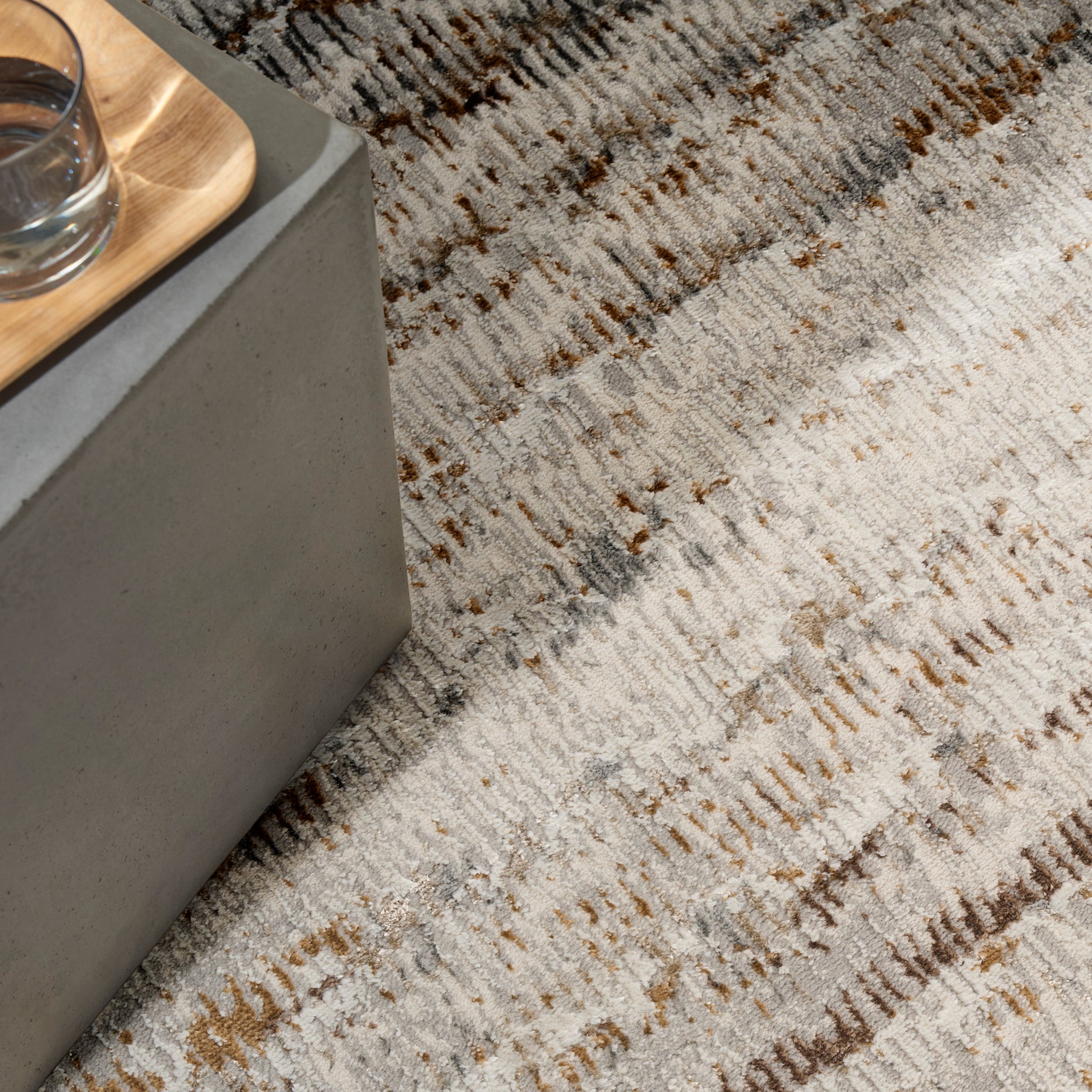 Close-up of a textured beige-gray rug with brown streaks beside a concrete edge and wooden tray with a glass.