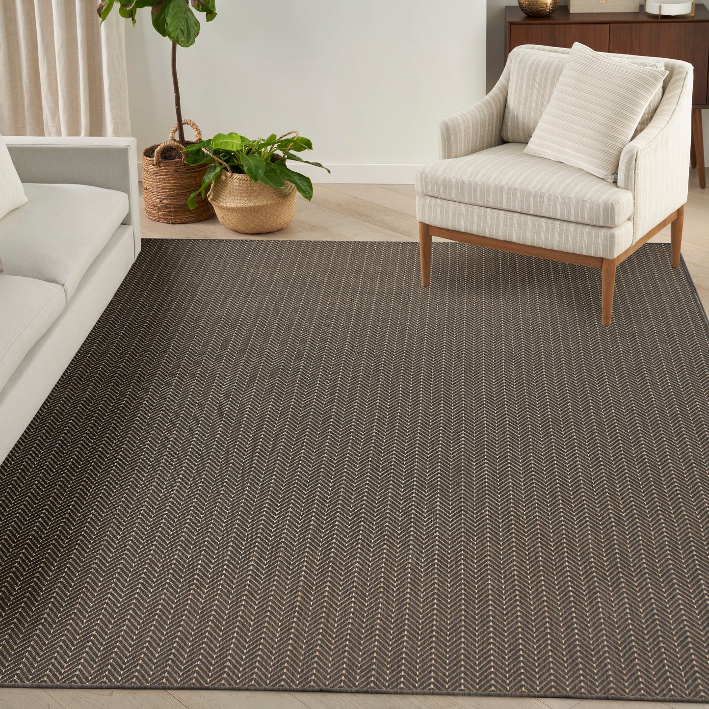 Dark herringbone rug anchors a light living room with cream sofa, striped beige armchair and potted plants.