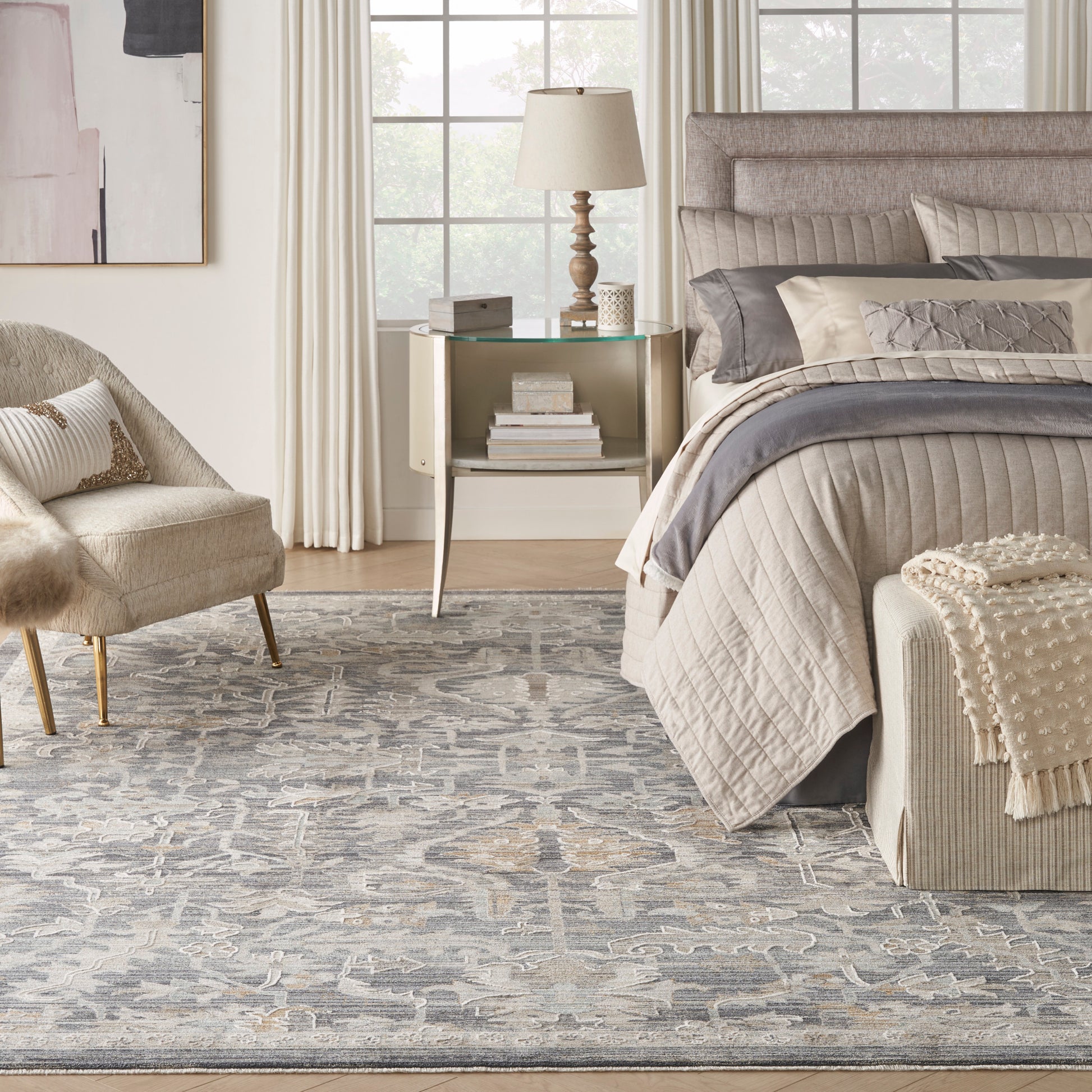 Neutral beige bedroom with quilted taupe bedding, tufted headboard, glass-top nightstand, & gray-blue rug.