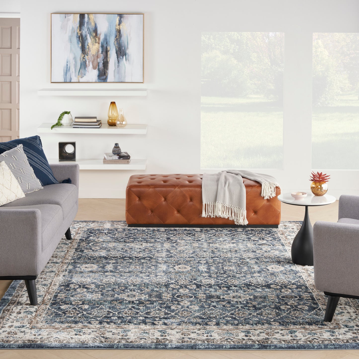 Modern living room with blue-gray patterned rug, gray sofas, tan tufted ottoman, white shelves, and abstract wall art.