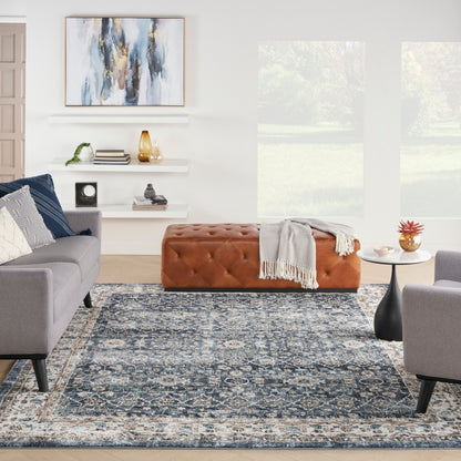 Modern living room with blue-gray patterned rug, gray sofas, tan tufted ottoman, white shelves, and abstract wall art.