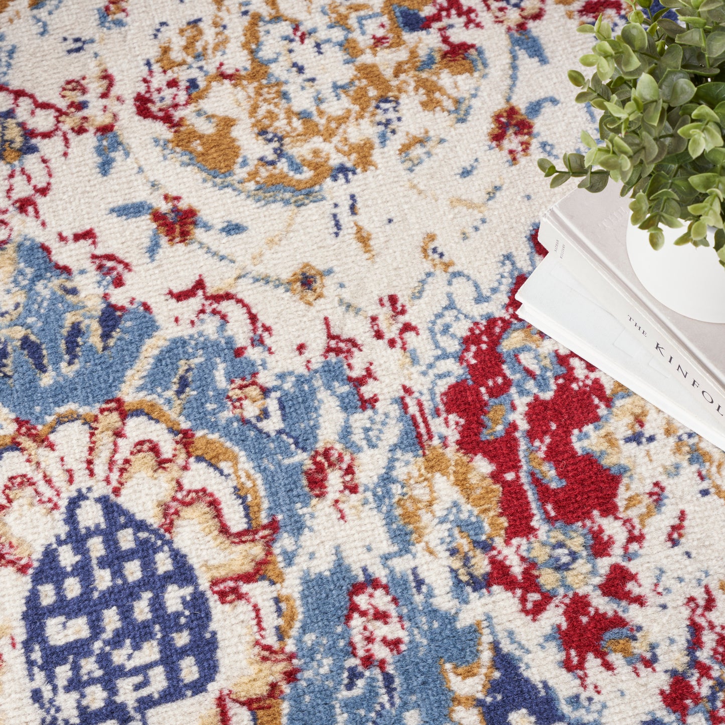Close-up of a plush oriental rug with cream base and blue, red, mustard floral motifs; plant and magazines at edge.