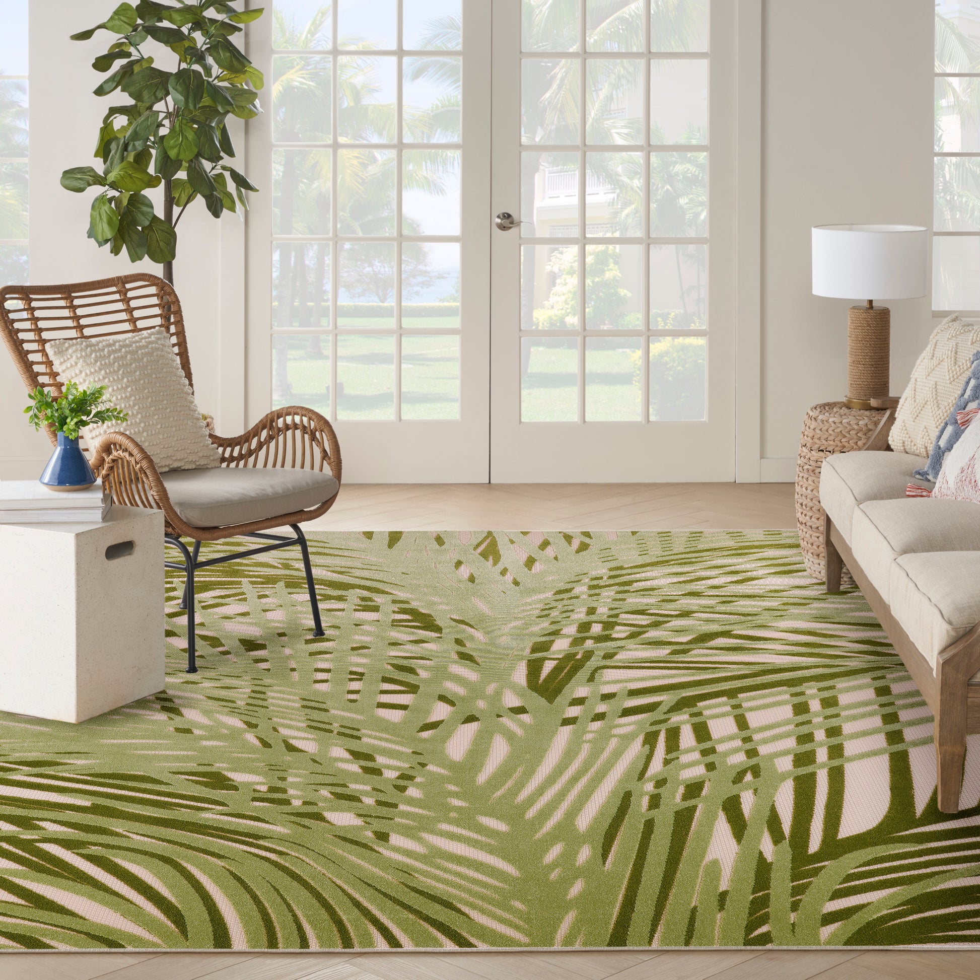 Bright living room with palm-leaf rug, wicker chair, beige sofa, tall plant, glass doors, tropical-inspired decor.