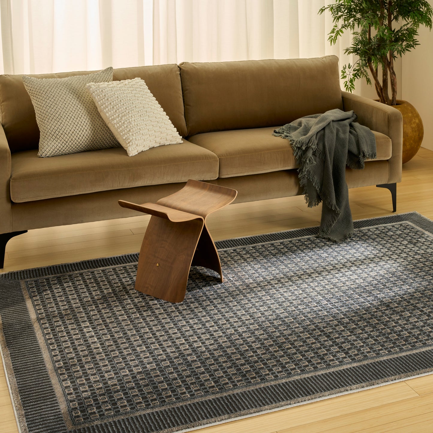 Tan fabric sofa with two textured cushions, gray throw, wooden curved stool, blue-gray geometric rug and a potted plant.