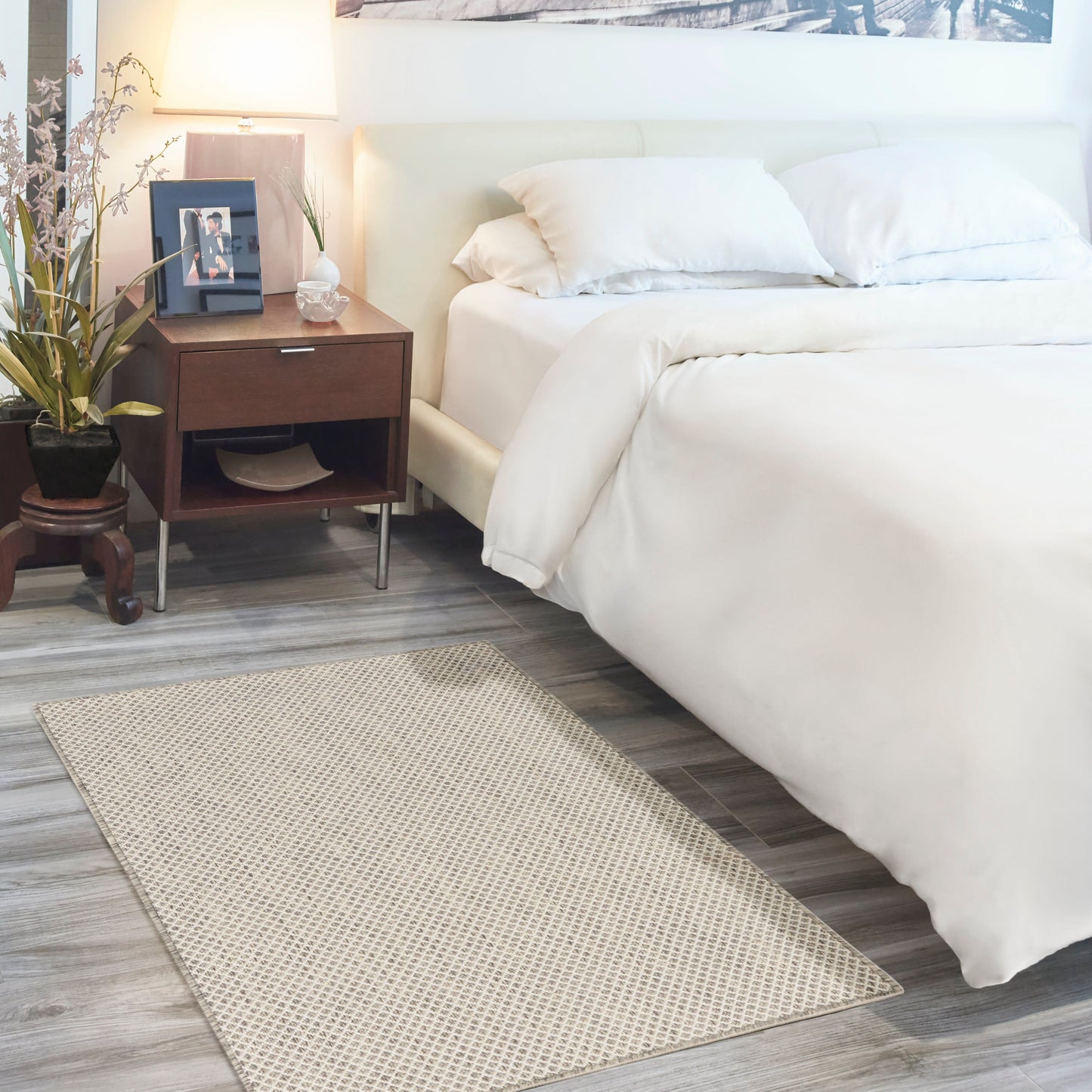 White bed with matching duvet, wooden nightstand, lamp, framed photo, potted plant, beige textured rug on gray flooring.