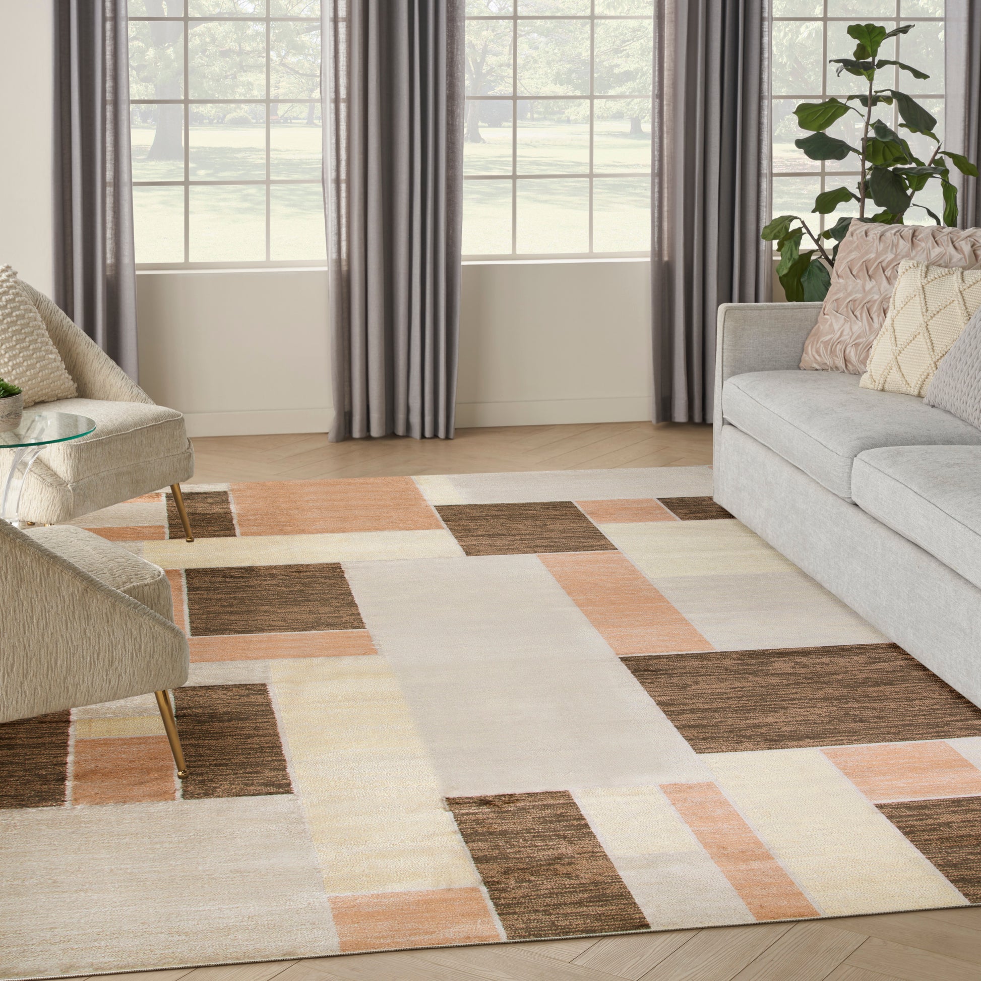 Living room with a color-block rug in beige, cream, brown & peach, gray sofa, beige armchair, plant, & large windows.