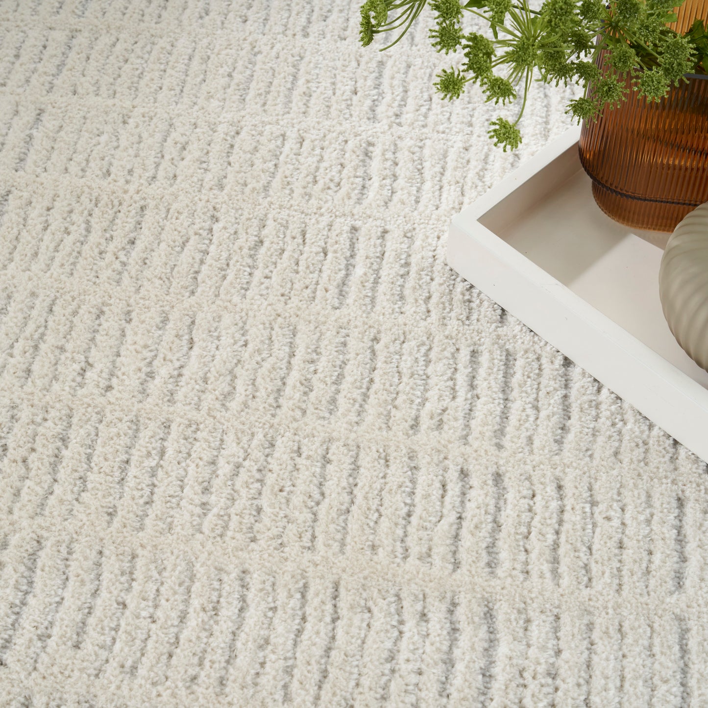 Cream textured rug with a white rectangular tray holding an amber glass vase and green foliage.