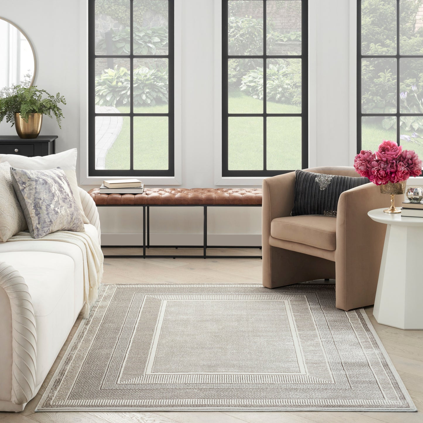 Light gray geometric rug in a bright living room with beige sofa, brown leather bench, black window frames, and pink flowers.