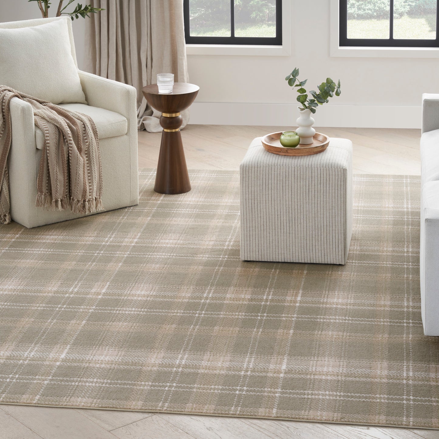 Cozy neutral living room with beige plaid rug, cream sofa, midcentury wooden side table, striped fabric ottoman topped by a wooden tray with greenery.