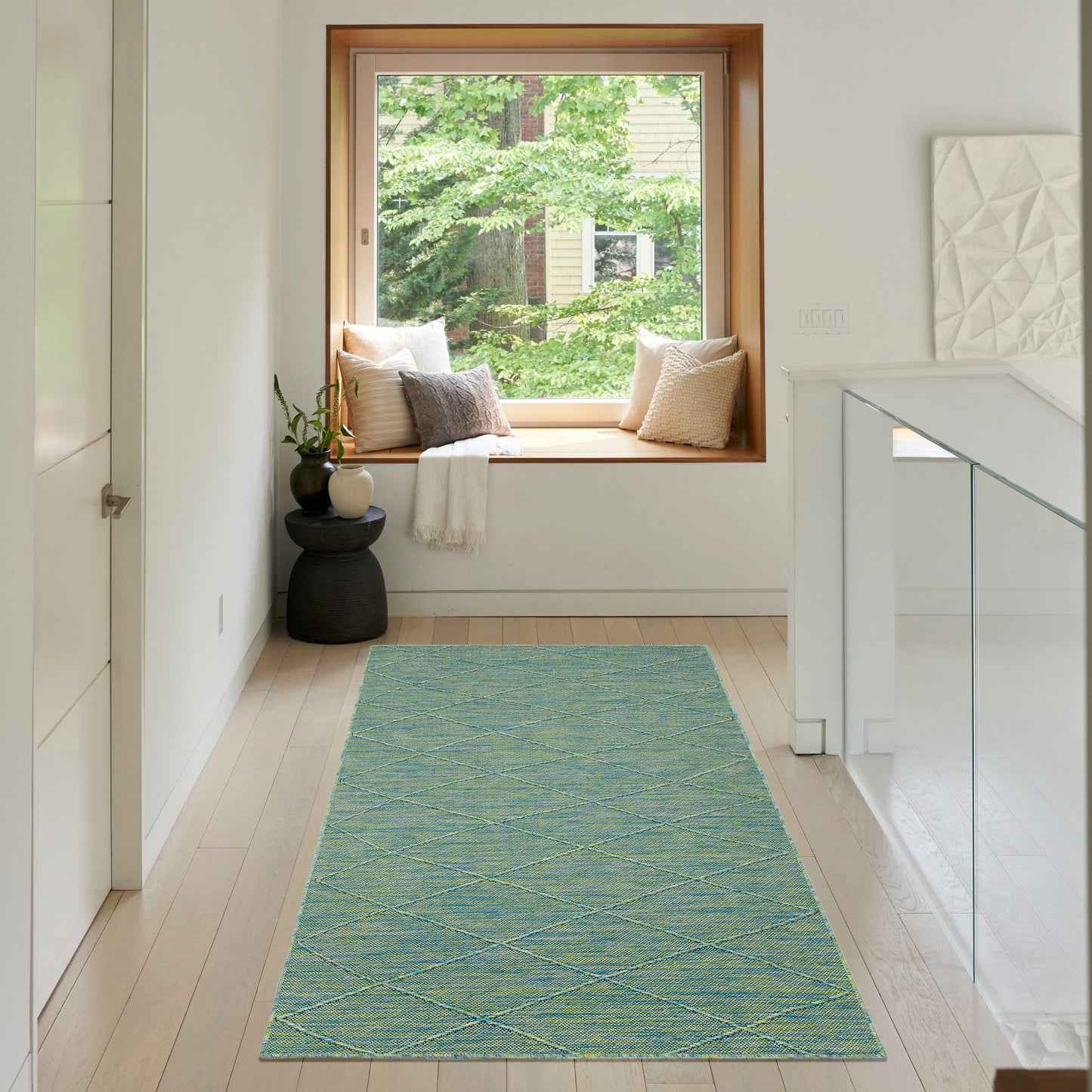 Turquoise diamond-pattern rug runner on light wood floor in a white hallway with window seat, cushions and glass railing.