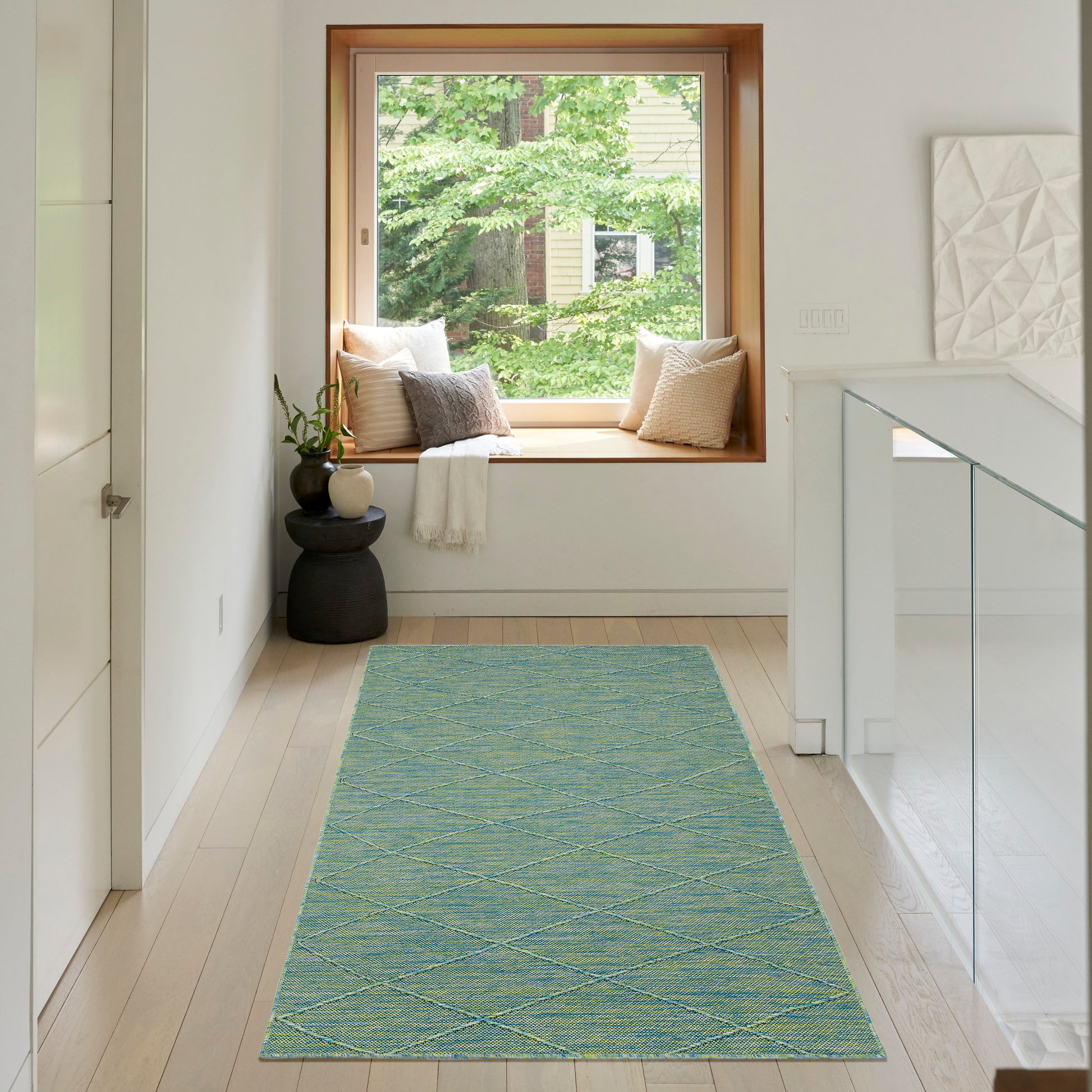 Turquoise diamond-pattern rug runner on light wood floor in a white hallway with window seat, cushions and glass railing.