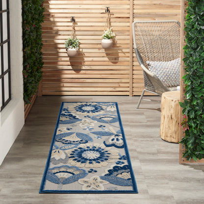 Blue floral outdoor runner rug on beige background, laid on a wooden deck beside a wicker chair and hanging planters.