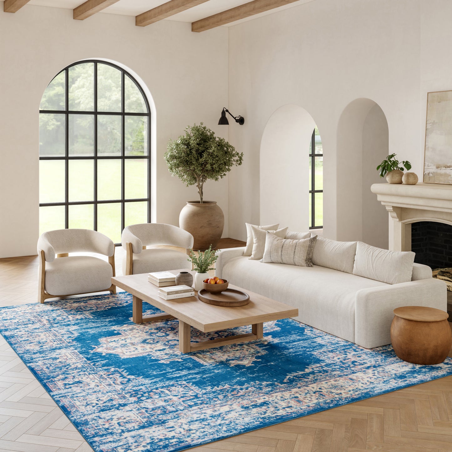 Bright living room with arched window, beige sofa, cream chairs, wooden coffee table, blue rug, potted tree, & fireplace.