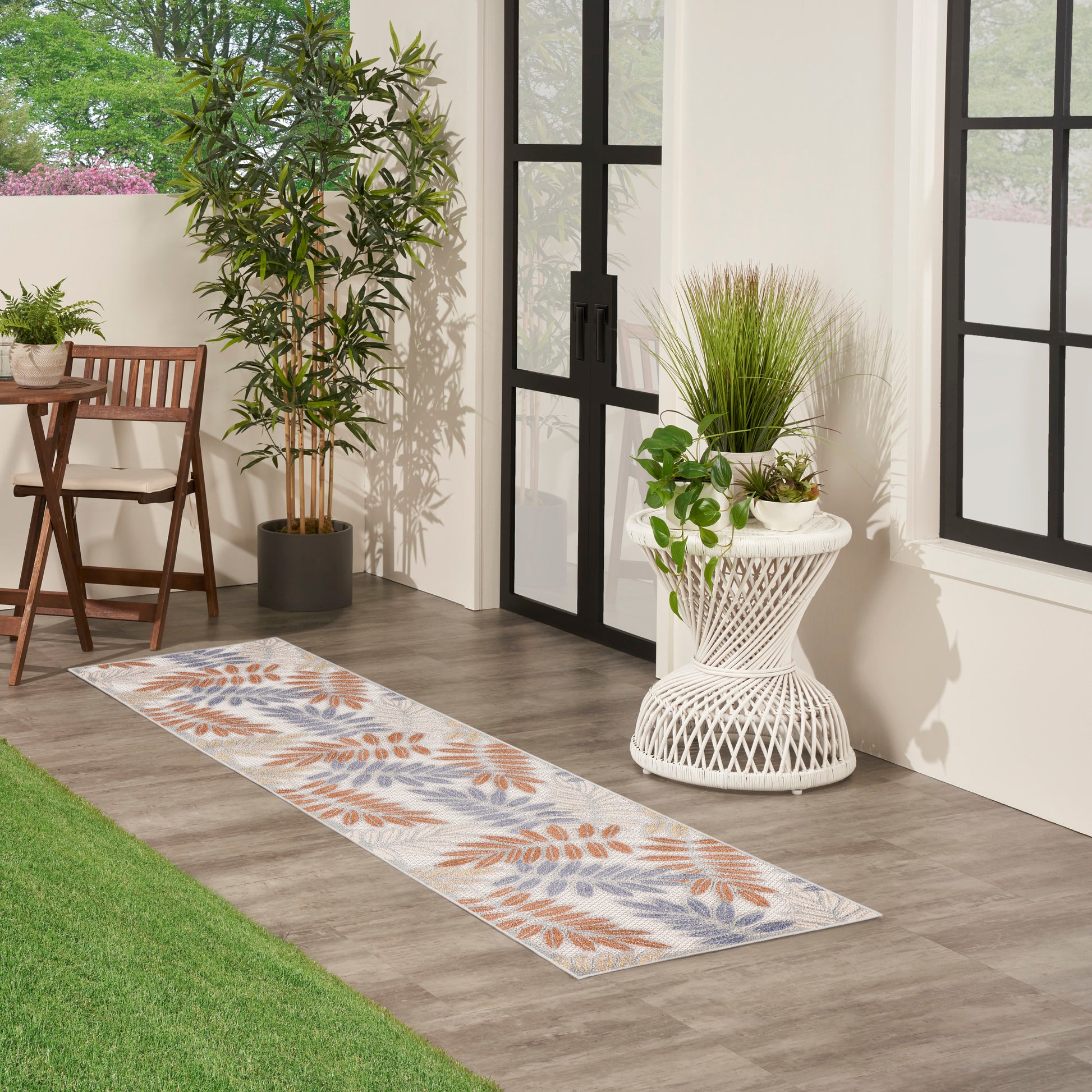 Long runner rug with orange and blue botanical leaves on light background beside potted plants and a black-framed glass door.