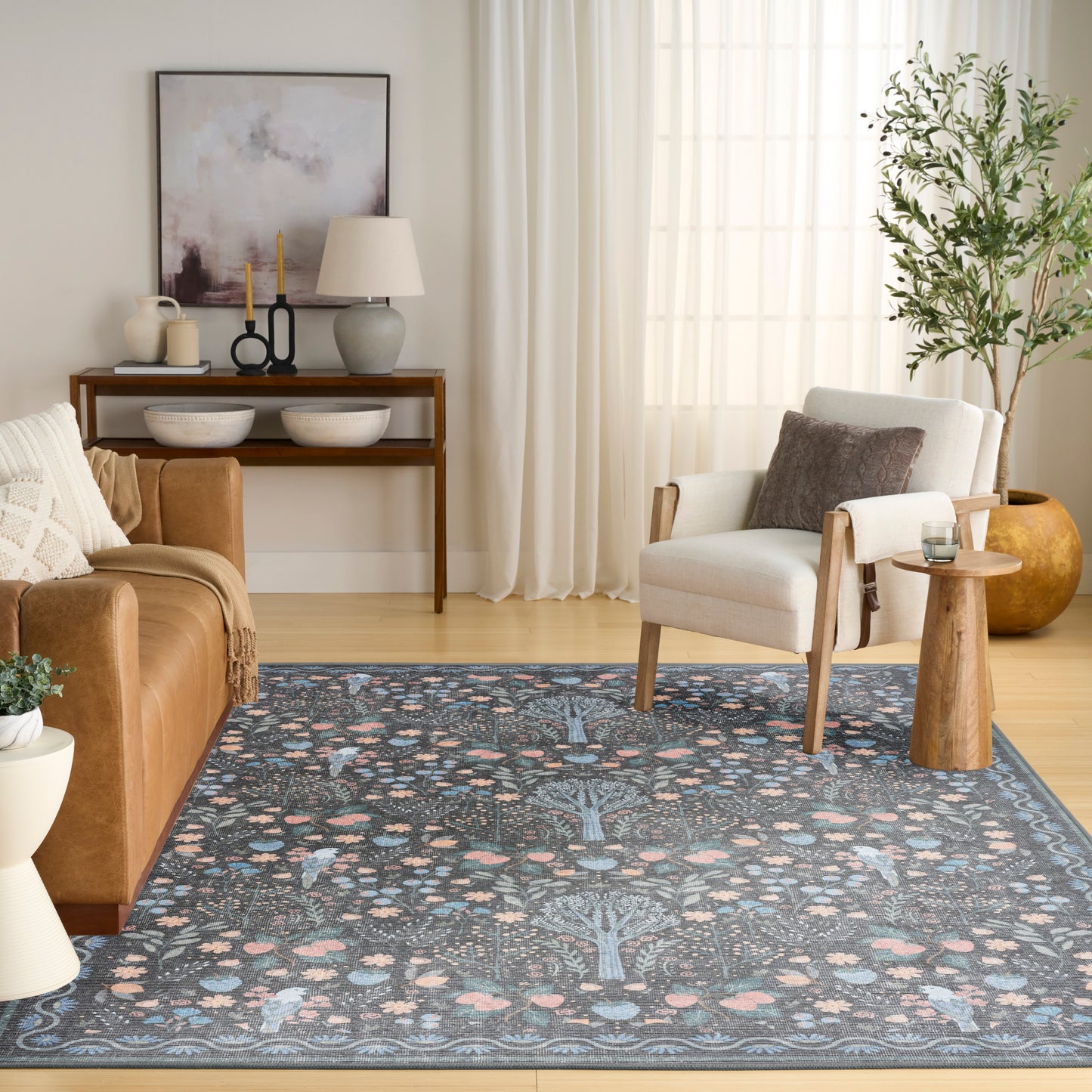 Light-filled living room with blue floral area rug, tan leather sofa, white armchair, wooden side tables, and lush potted plant.