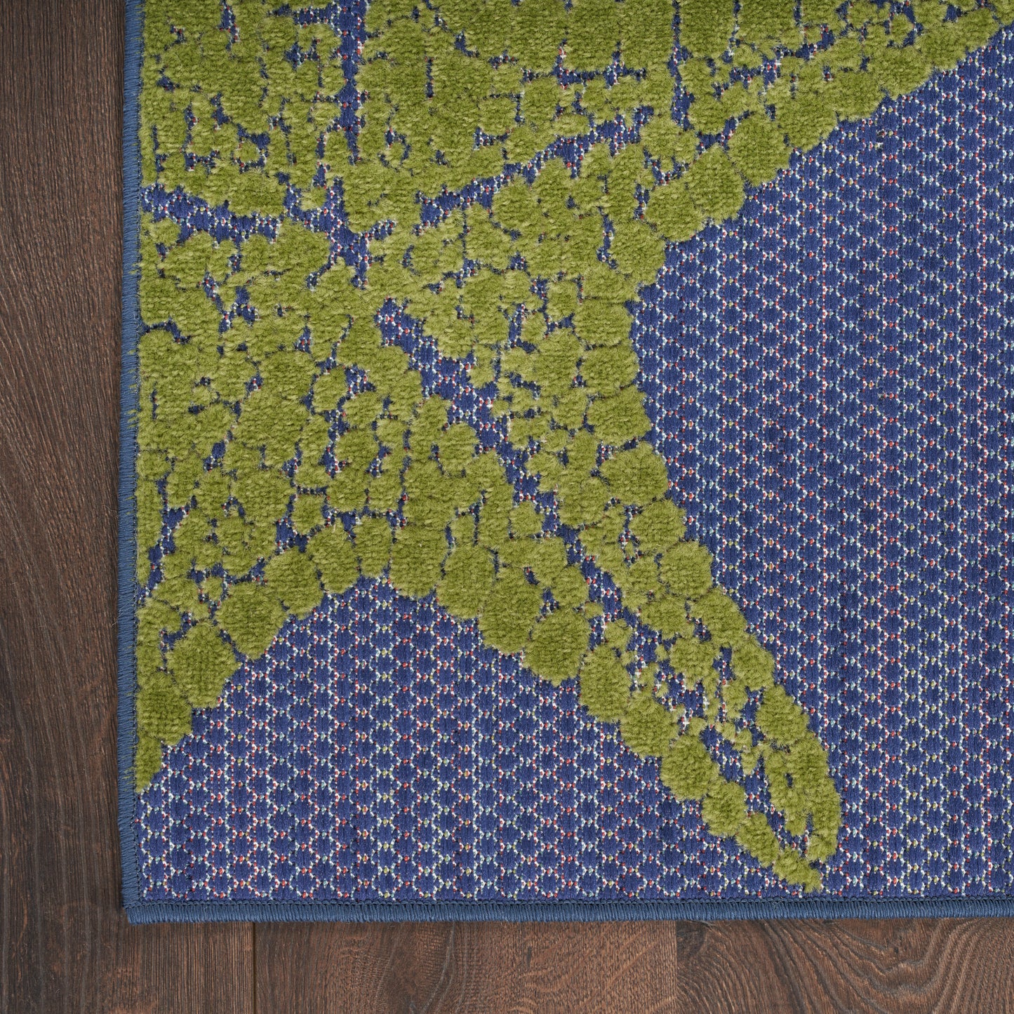 Close-up of an outdoor rug corner with a lime-green botanical motif on a blue speckled field; dark blue border, wood deck nearby.