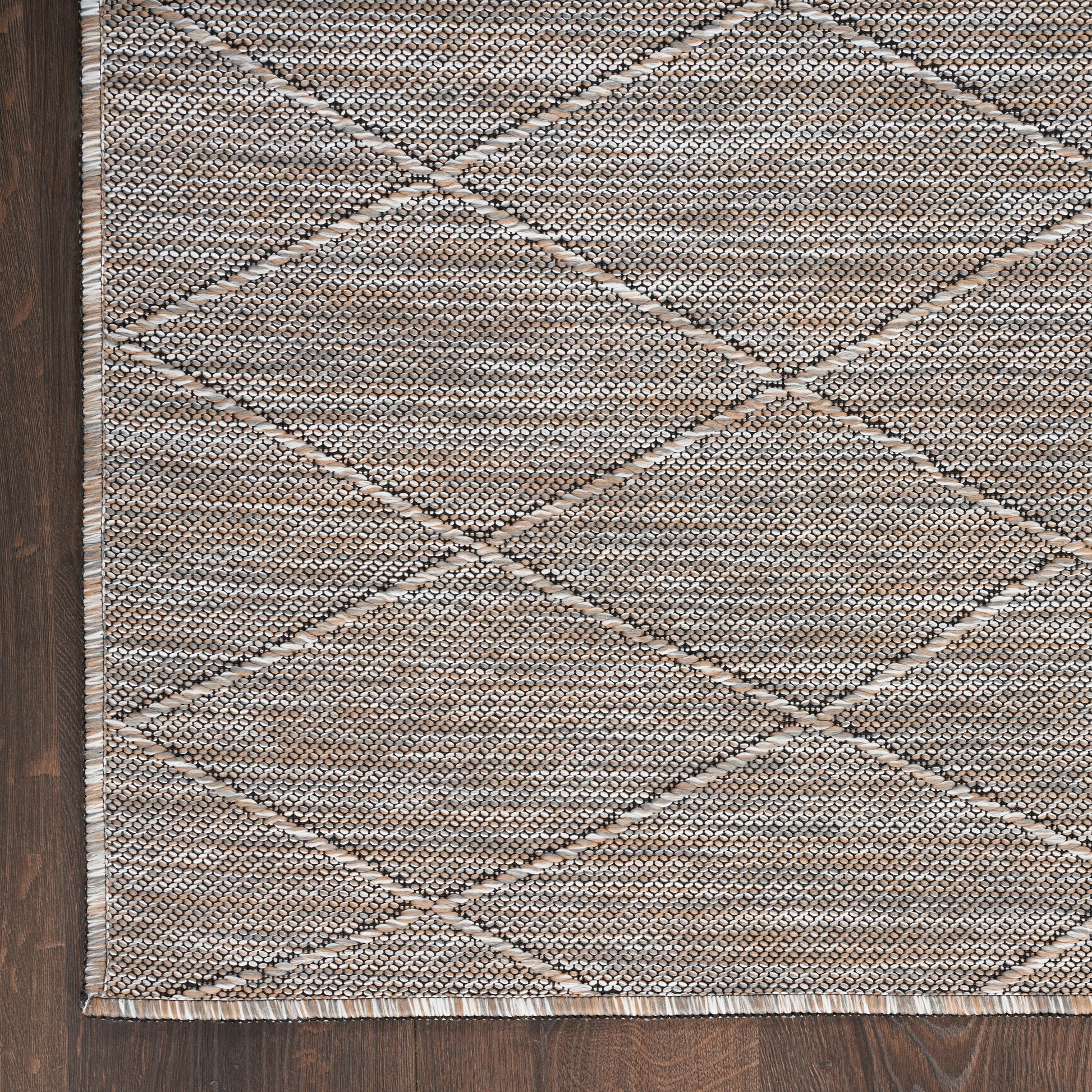 Beige woven rug featuring a geometric diamond pattern with fringe along the edge on dark wood floor.