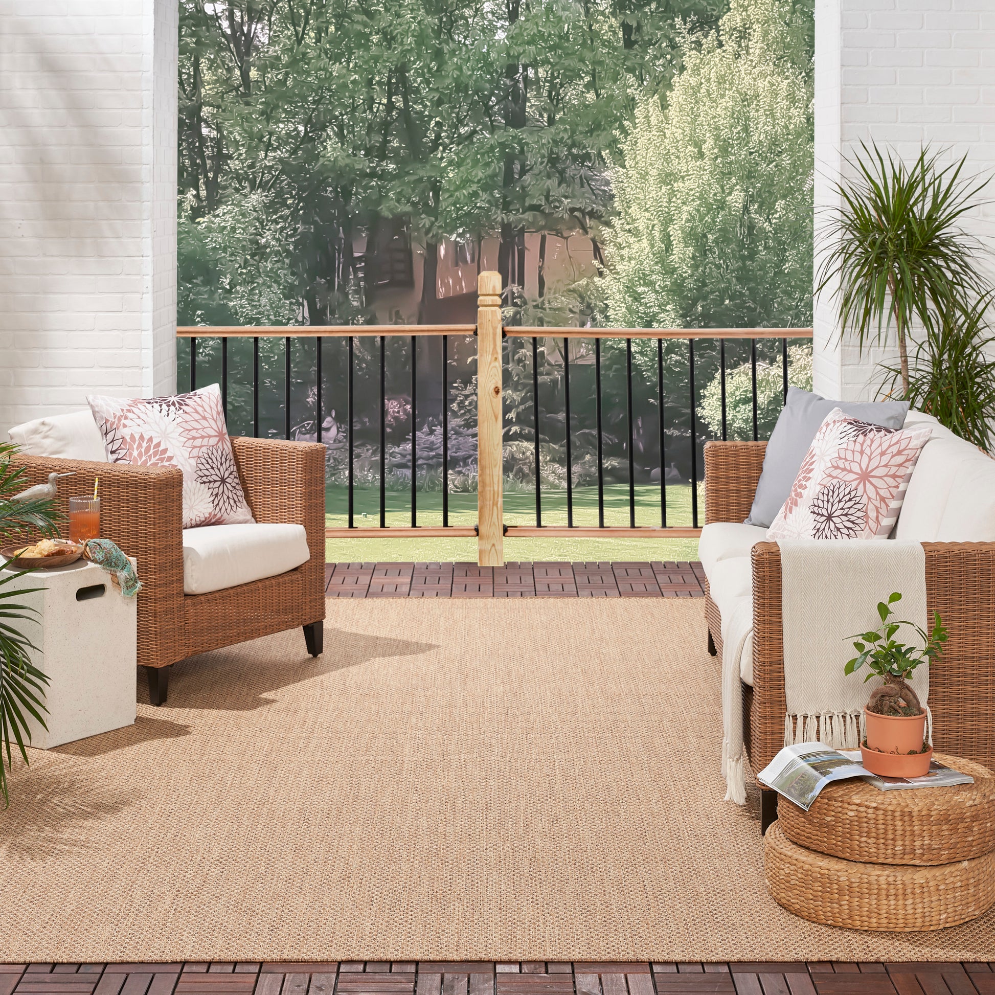 Cozy outdoor wicker seating on a deck with a beige rug, cushions & pillows, potted plant, & garden beyond railing.
