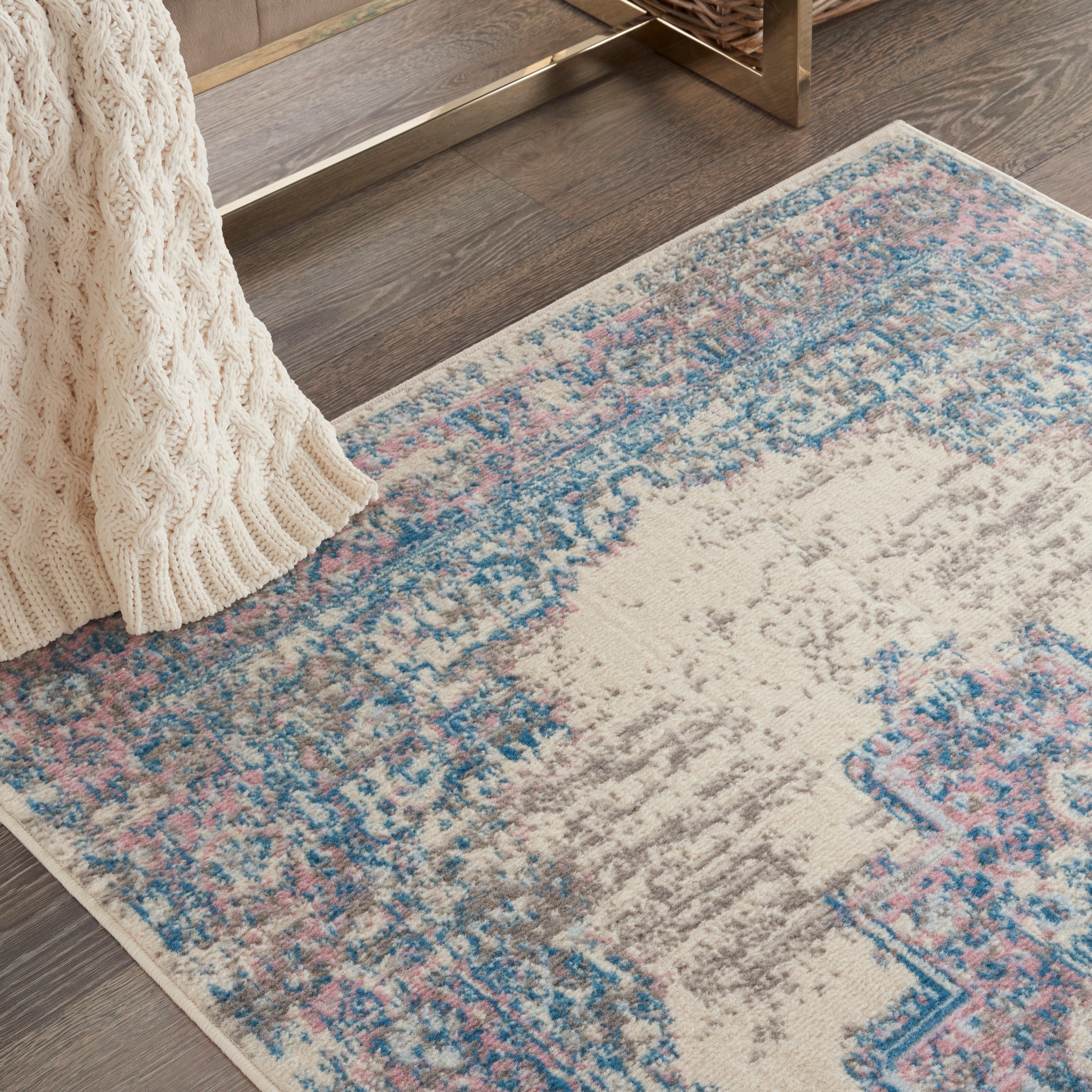 Distressed cream and blue oriental rug with pink accents on dark wood floor, beside a chunky beige knit throw.