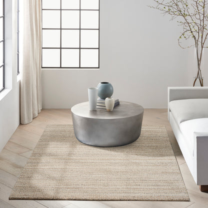 Beige woven rug in a bright minimalist living room, with a round coffee table, ceramic vases and books visible.