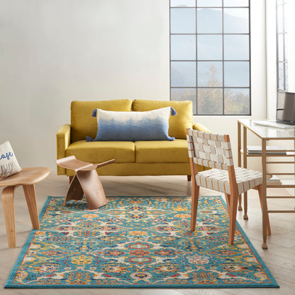 Mustard yellow fabric sofa with blue tassel pillow, teak chair, curved wooden stool, glass desk, and teal patterned rug