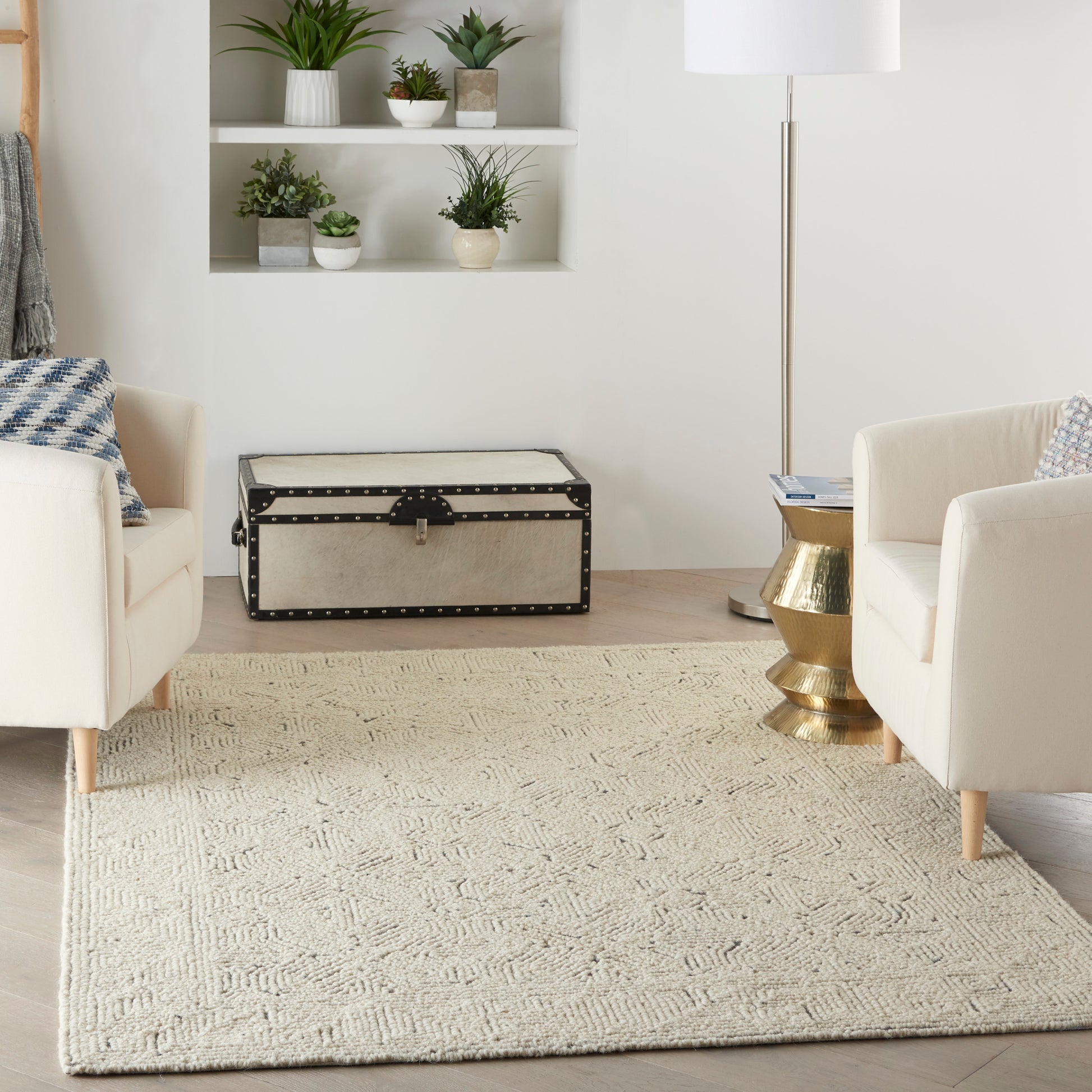 Cream upholstered chairs, beige woven rug, vintage studded trunk, brass side table, and potted plants in a living room.