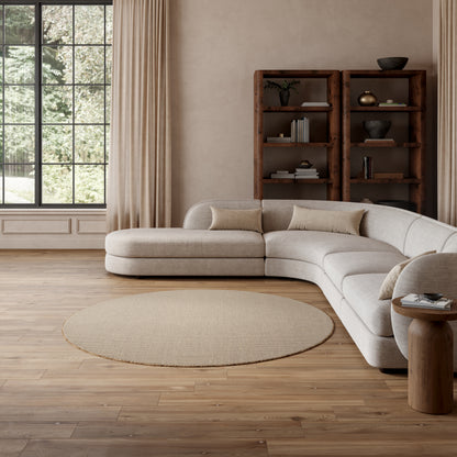 Beige L-shaped fabric sofa with cushions, circular beige rug, wood floor, tall bookshelf, airy living room with grid window.