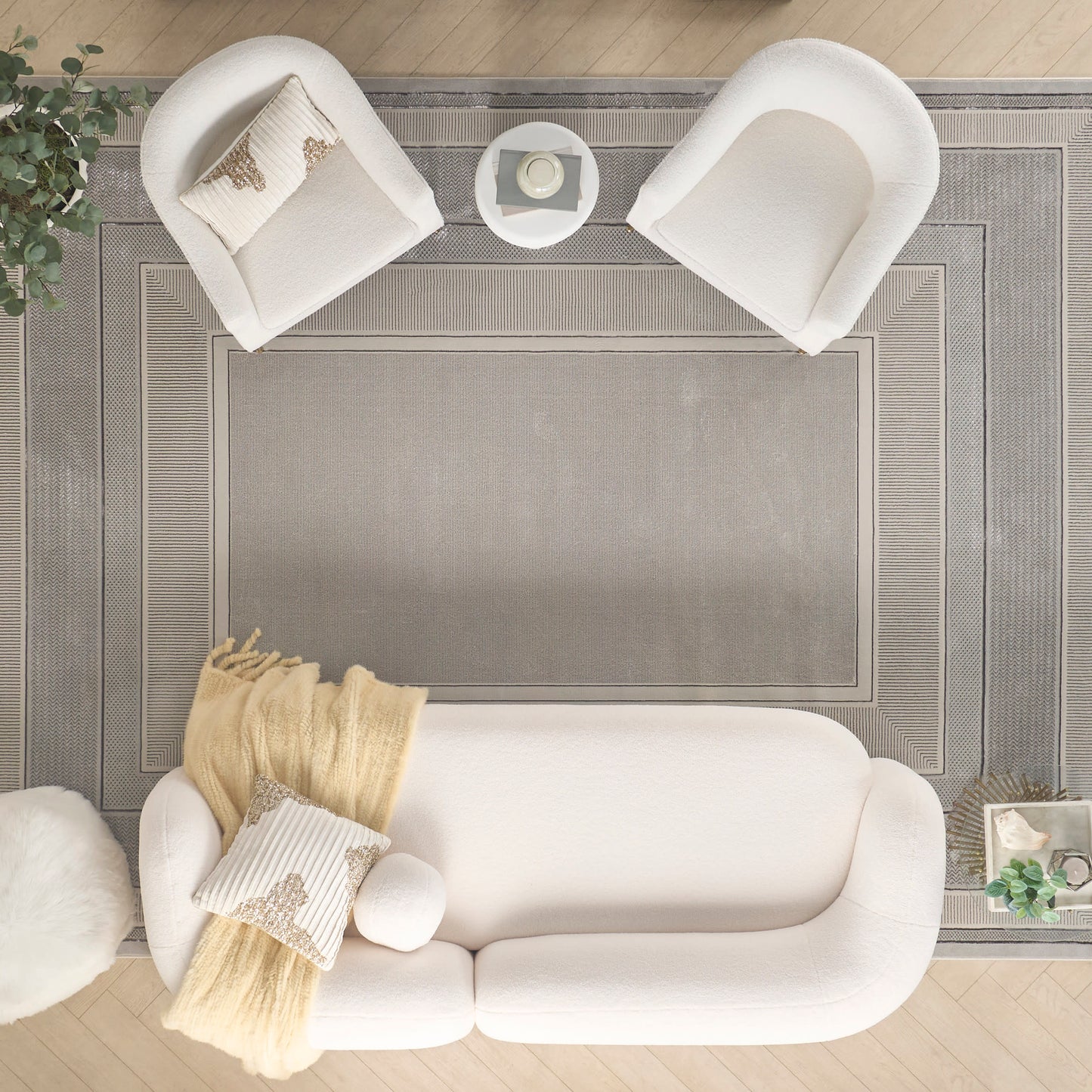 Top-down view of a white living room with a plush sofa, two curved chairs, a small round table, and a gray bordered rug.