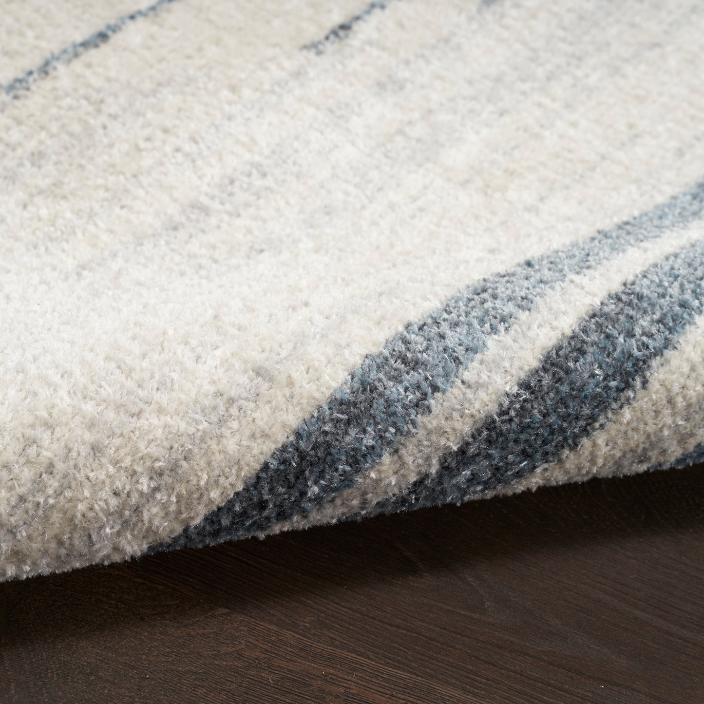 Cream shag rug with blue-gray stripes, folded edge on dark wood floor.