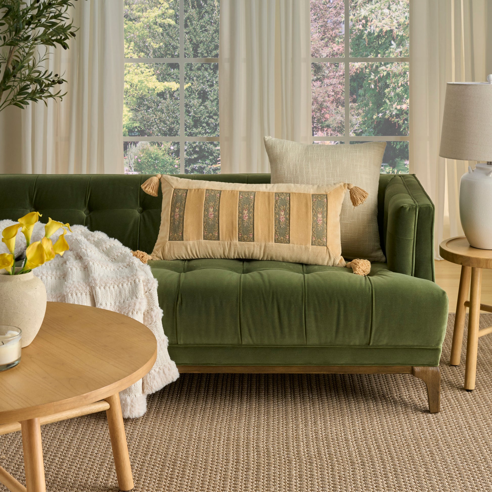 Sage green rug in a living room setting, complemented by a beige tassel pillow, round wooden coffee table, cream throw, potted plant, and windows.