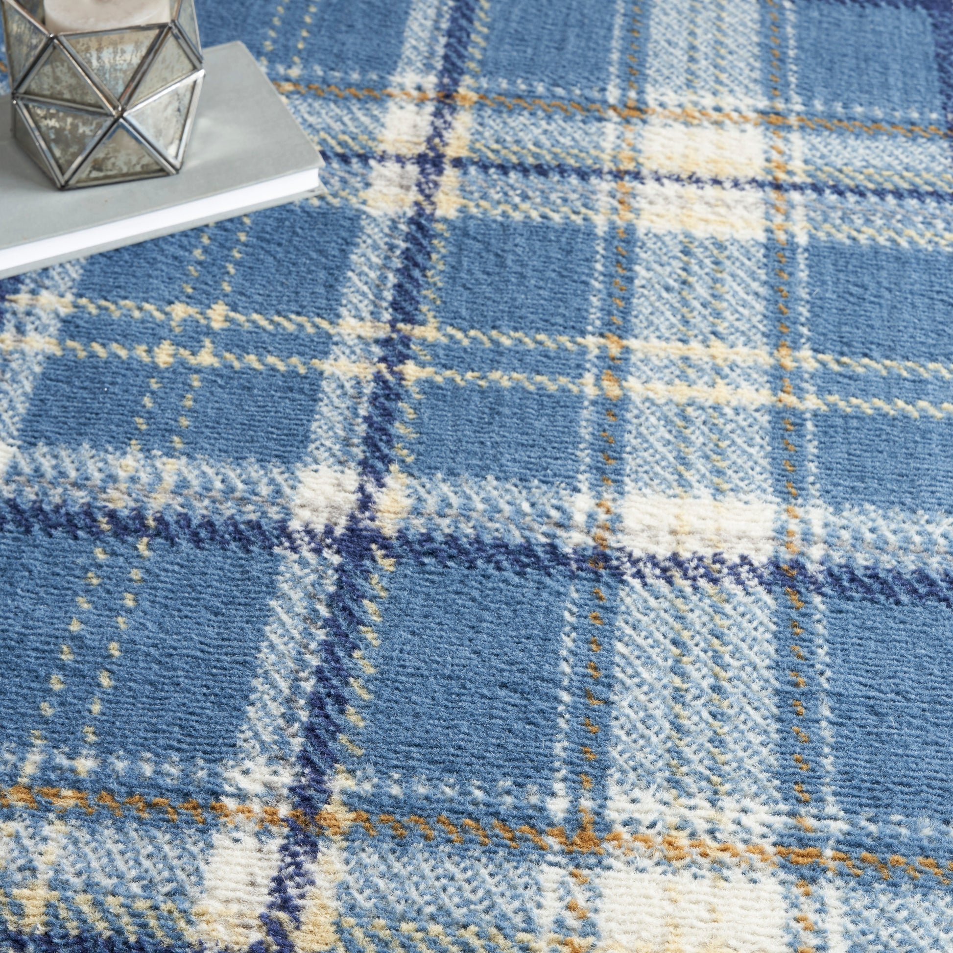 Blue plaid wool rug with cream and rust lines; cozy decor setup with a geometric metal candle holder on books.