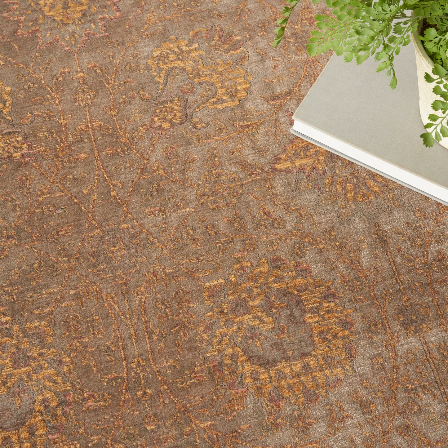 Earthy brown floral rug with gold and amber damask pattern; top-right corner shows a gray book and green fern.