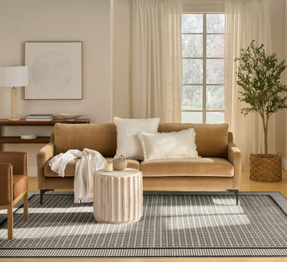 Warm tan sofa with white pillows, round ribbed wood coffee table, neutral rug, cozy throw, and a potted tree.
