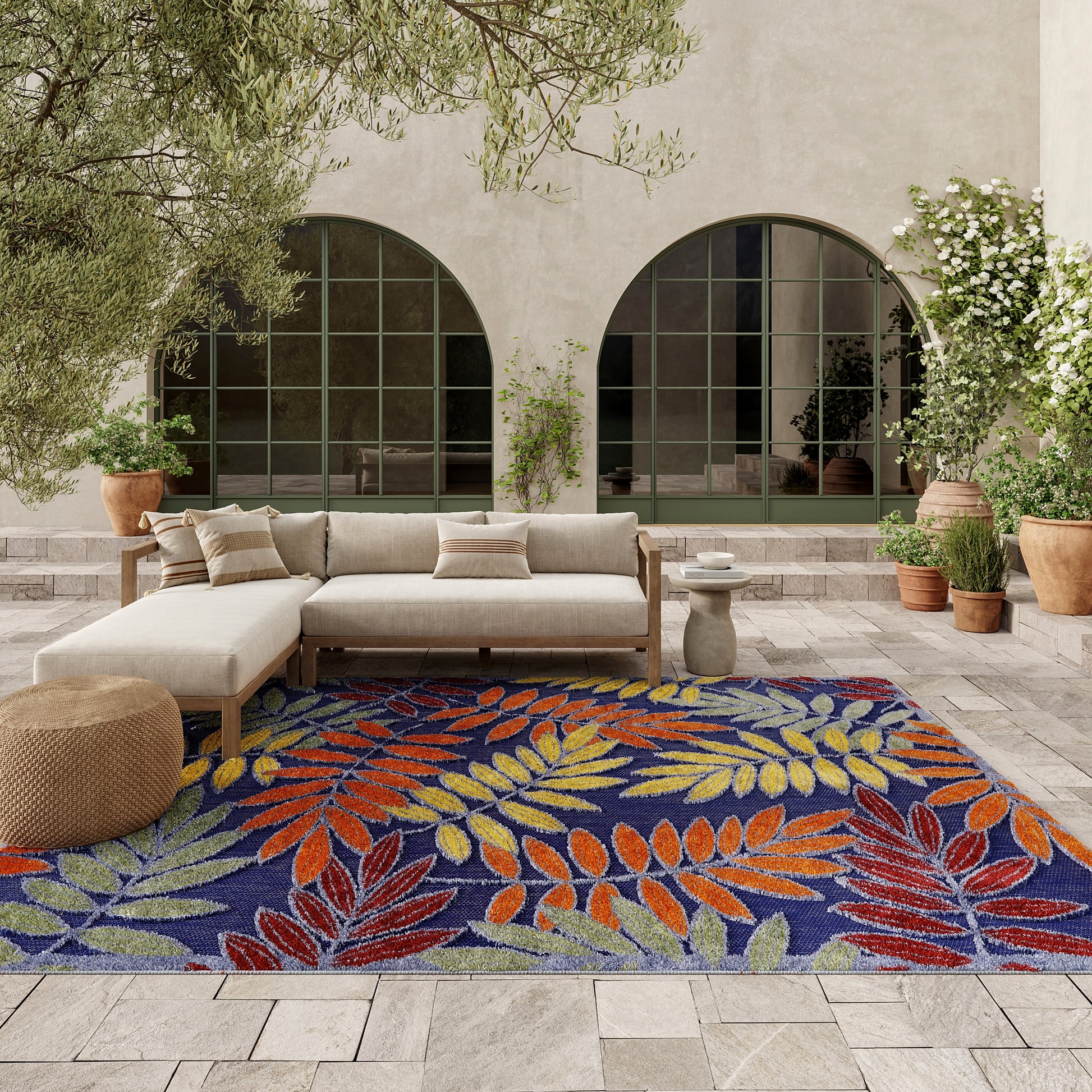 Outdoor patio with beige sectional sofa on a vibrant leaf-pattern rug in blue, orange and yellow; potted plants and arched windows.