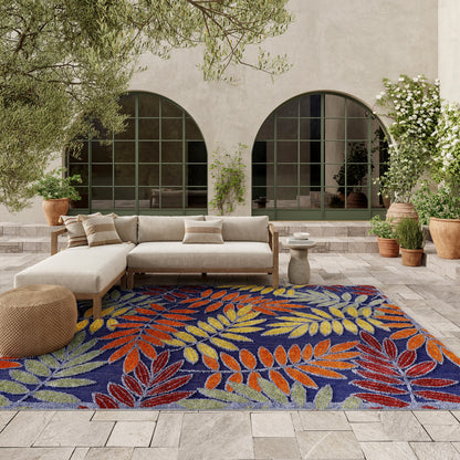 Outdoor patio with beige sectional sofa on a vibrant leaf-pattern rug in blue, orange and yellow; potted plants and arched windows.