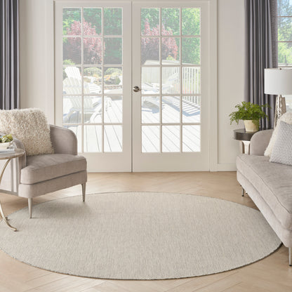 Bright living room features a round beige woven rug, gray sofas, light wood floors, and French doors opening to patio.