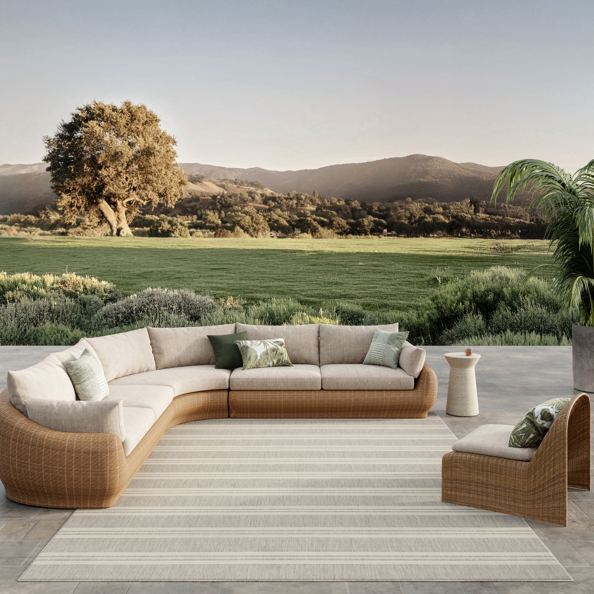 Outdoor beige wicker sectional sofa with cream cushions and green pillows on a striped rug, set against a scenic landscape.