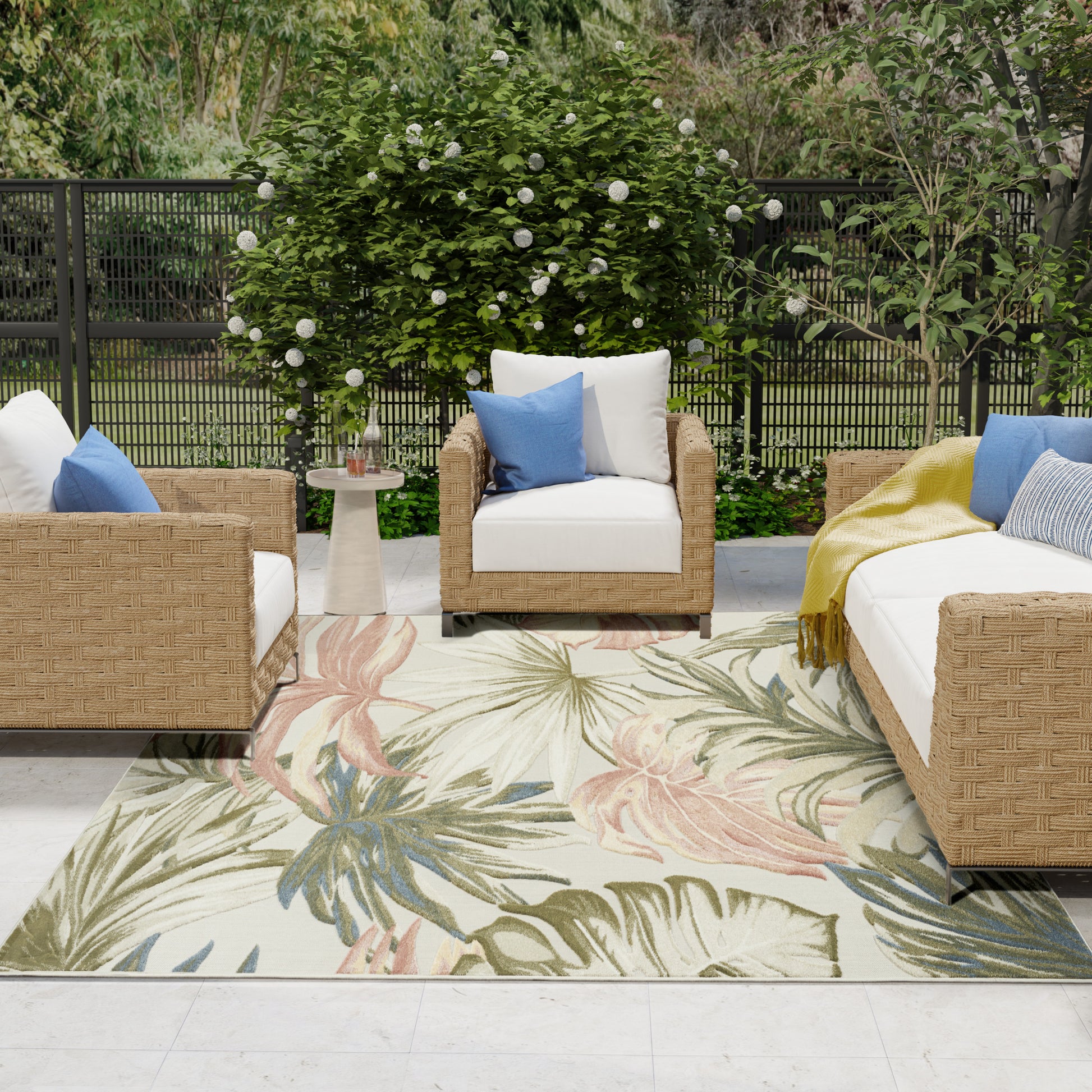 Outdoor wicker patio furniture set with white cushions, blue pillows, tropical leaf rug, and lush garden backdrop.
