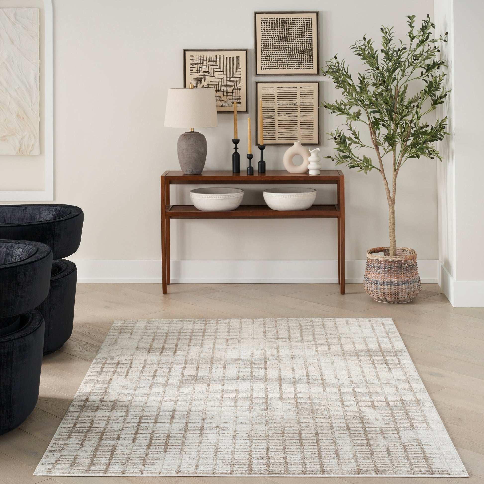 Neutral living room with wooden console, ceramic lamp, framed abstract art, bowls, tall plant in woven basket, beige rug.