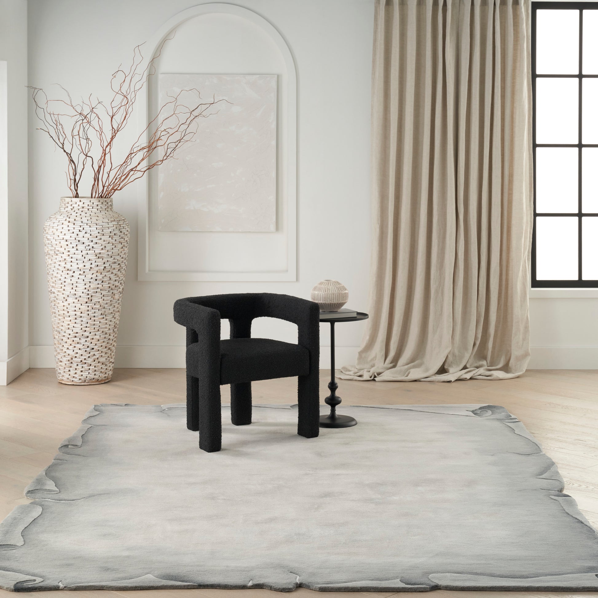 Minimalist living room: black curved lounge chair, round side table, speckled vase with branches, pale rug, beige curtains.