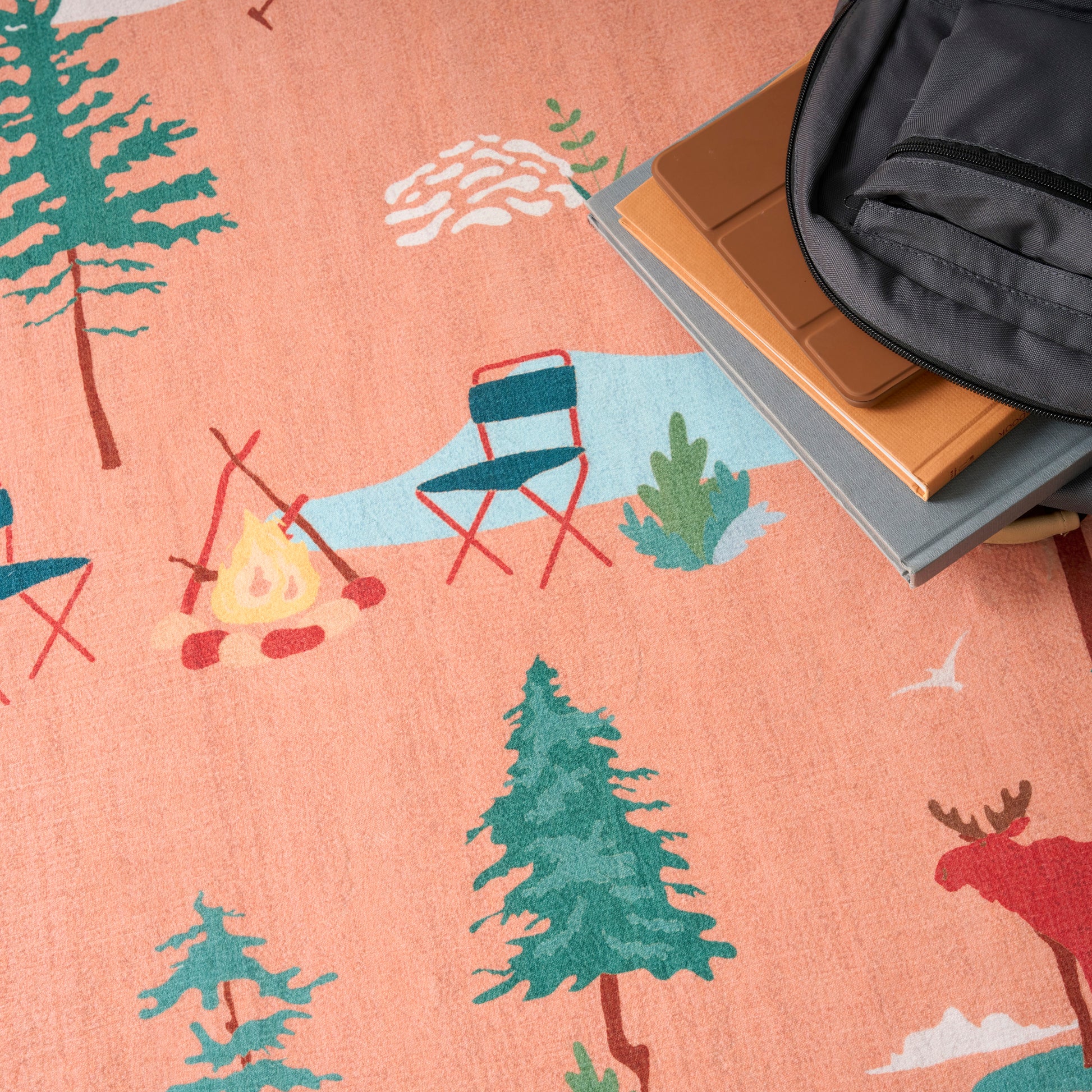 Peach-colored camping scene rug with teal trees, campfire, blue chairs; backpack and books along the edge.