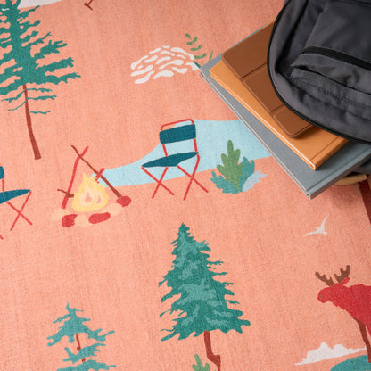 Peach-colored camping scene rug with teal trees, campfire, blue chairs; backpack and books along the edge.